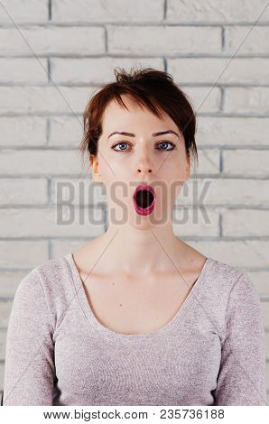 A Pretty Girl With Surprised Face, Big Blue Eyes, Brows Up, With White Brick Wall In The Background.