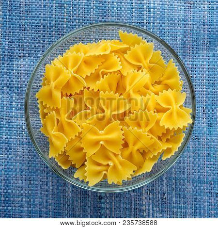 Farfalle Raw Pasta In A Glass Bowl On A Blue Knitted Background In The Center. Close-up With The Top