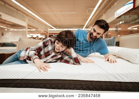 Handsome Bearded Father With Young Son Is Testing Mattress For Softness. Testing Softness Of Mattres