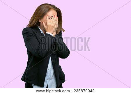 Middle age business woman smiling having shy look peeking through her fingers, covering face with hands looking confusedly broadly