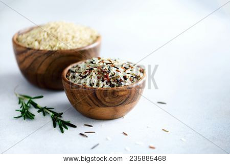 Raw Organic Brown Rice In Wooden Bowl And Rosemary On Light Concrete Background. Food Ingredients. C