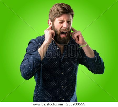 Young hipster man with big beard covering ears ignoring annoying loud noise, plugs ears to avoid hearing sound. Noisy music is a problem. over green background