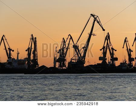 The Sea And Dark Silhouettes Of Cargo Cranes In The Seaport On The Bright Orange Sky At Sunset