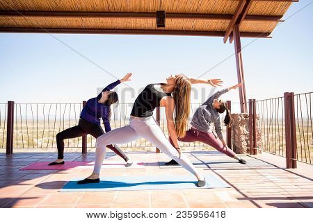 Three Hispanic Women Standing And Stretching Backward In Reverse Warrior Pose At Yoga Class