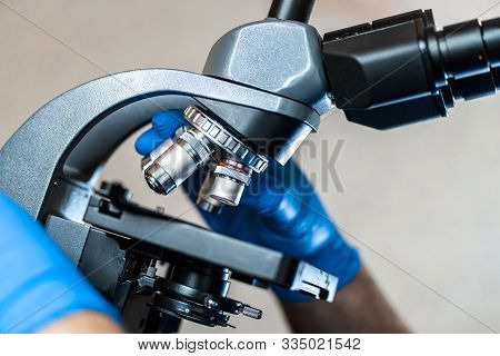 Male Laboratory Assistant Examining Biomaterial Samples In A Microscope. Cllose Up Hands In Blue Rub
