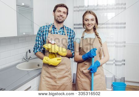 Young Man And Woman Posing In Aprons With Household Stuff Amidst A Light Kitchen