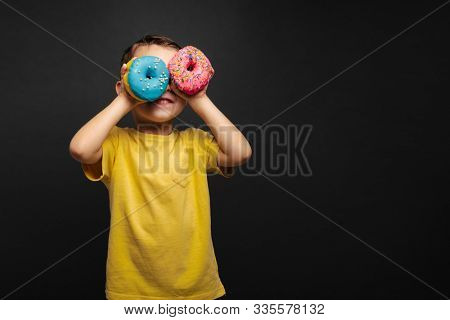 Happy cute boy is having fun played with donuts on black background wall. Bright photo of a boy. Colored donuts