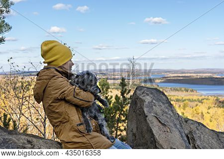 Young Woman In Brown Jacket Sitting With Fluufy Gray Dog In Arms On Rock And Looking At Beautiful Au