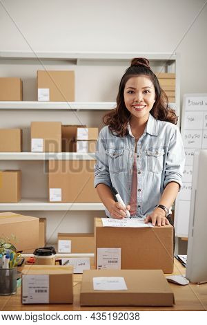 Portrait Of Smiling Young Asian Woman In Denim Jacket Standing At Table With Parcels And Filling Con