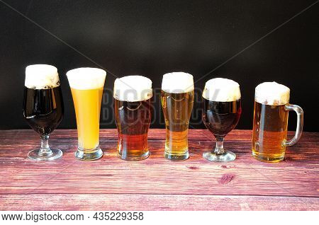 Six Different Glasses Of Beer With Six Different Types Of Beer With Froth Stand In A Row On A Wooden