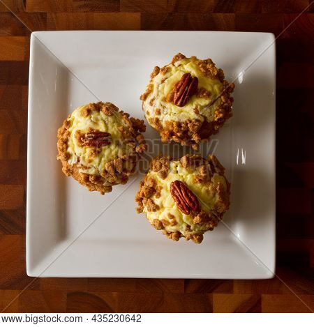 Birds Eye View Of Three Orange Muffins Topped With Frosting And A Pecan On A White Plate On A Table