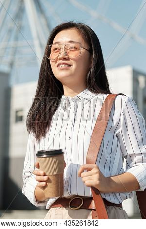 Happy young Chinese businesswoma with drink standing against amusement park