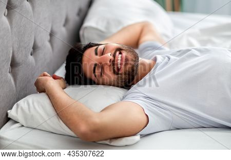 Happy Wellslept Arab Man Waking Up In Morning, Stretching And Smiling With Closed Eyes While Lying I