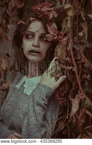 A sad lonely ghost girl with faded eyes and matted black hair stands among withered foliage and looks at the camera. Horror film, thriller. Halloween.