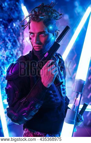 Courageous cyberpunk warrior in a protective costume poses with a gun in his hand standing among neon lamps. Game, virtual reality. 