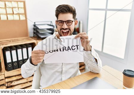 Young man with beard breaking contract paper at the office smiling and laughing hard out loud because funny crazy joke. 