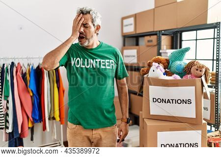 Middle age hispanic man wearing volunteer t shirt at donations stand yawning tired covering half face, eye and mouth with hand. face hurts in pain. 