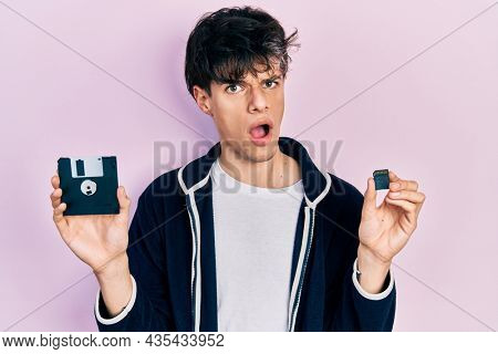 Handsome hipster young man holding floppy disk and sdxc card in shock face, looking skeptical and sarcastic, surprised with open mouth 