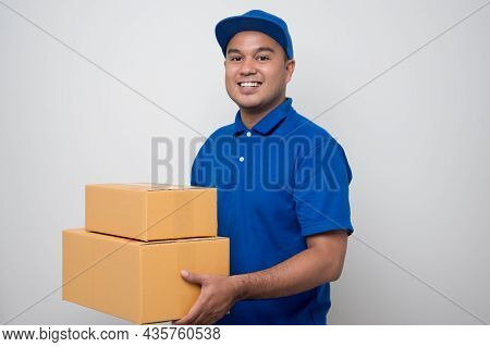Young Smiling Asian Delivery Man In Blue Uniform Holding Messenger Bag Showing Thumbs Up Sign Standi