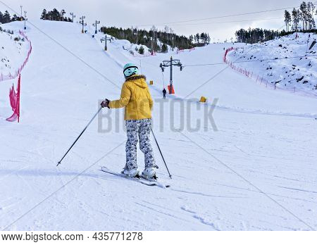Rear View Of Young Woman In Yellow Jacket And Ski Helmet Downhill Skiing On A Mountain Slope, Winter