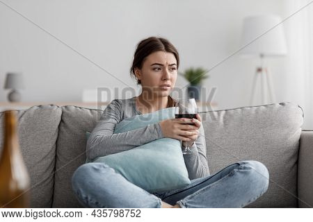 Upset Apathetic European Millennial Woman Sits On Sofa With Glass And Suffering From Depression And 