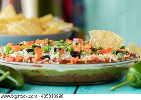 A 7 Layer Dip With One Tortilla Chip Dipped In And A Bowl Of Chips In Behind.