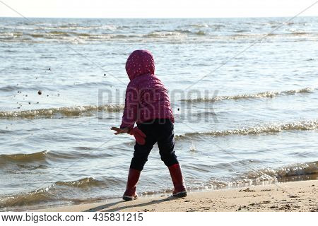 Child Girl Playing By The Sea In Autumn. A Girl In A Red Jacket In A Beautiful Autumn On The Seashor