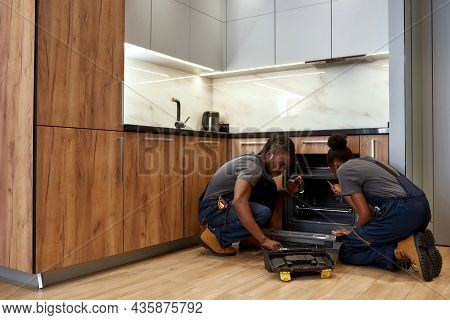Young African American Woman And Man, Repairpersons Kneeling In Front Of Opened Oven In Modern Kitch
