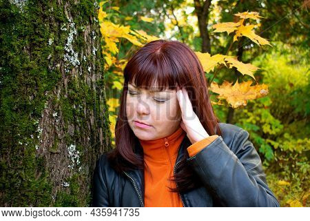 Red-haired Middle-aged Woman Is Having A Headache Or Migraine And Is Leaning On A Tree In The Park. 