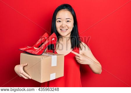Young chinese girl holding red high heel shoes and package smiling happy pointing with hand and finger 