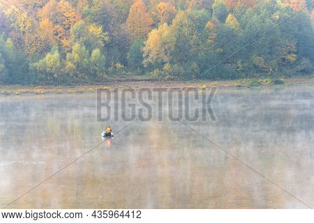 View Of A Fisherman In A Rubber Boat From The Back On The River And The Forest On The Opposite Bank.