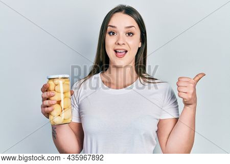 Young hispanic girl holding bottle with boiled potatoes pointing thumb up to the side smiling happy with open mouth 