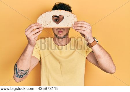 Young hispanic man holding bread with heart shape over eye puffing cheeks with funny face. mouth inflated with air, catching air. 