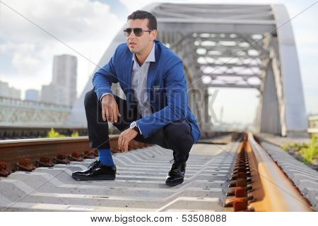 Stylish man in blue jacket and sunglasses squats on railroad near bridge and looks away.