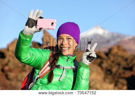Woman hiker taking selfie photo using smartphone while hiking in winter jacket and clothing enjoying outdoor activity. Woman hiker taking self-portrait picture with smart phone camera.