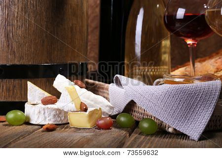 Supper consisting of Camembert and Brie cheese, wine and grapes on napkin in basket and wine barrel on wooden table on brown background