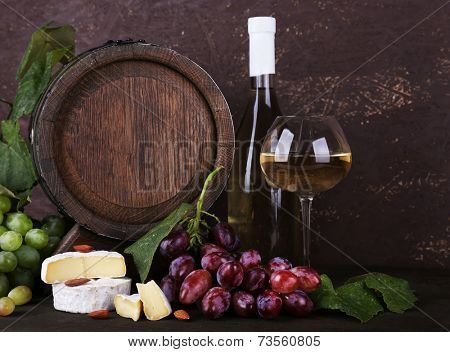 Wine in bottle and in goblet, Camembert and brie cheese, grapes and wooden barrel on wooden table on wooden background