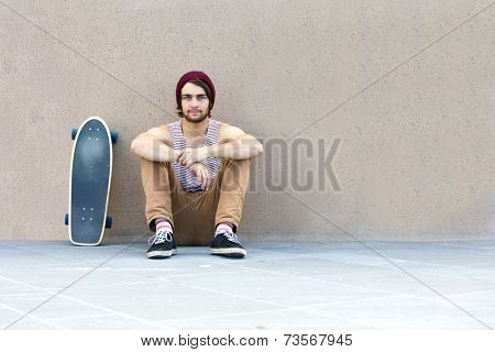Handsome skateboarder hanging around, sitting against a granite wall, with his skateboard next to him.