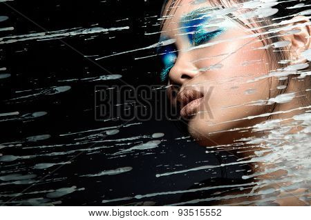 Beautiful Asian girl with bright blue make-up behind glass and drops of wax. Beauty face.