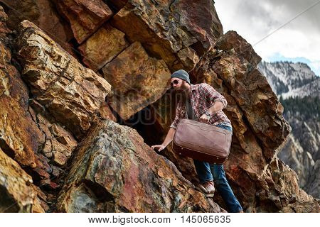 bearded hipster underestimates difficulty of carrying suitcase up a mountain shot with selective focus