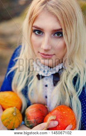 A woman dressed in a blue vintage dress sits in the autumn park. She holds a small bright pumpkins.