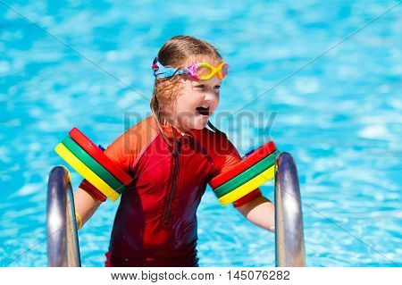Happy laughing little girl playing in outdoor swimming pool on a hot summer day in sun protection rash guard. Kid in goggles and UV safe bathing suit learning to swim with colorful floaties.