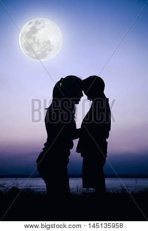 Silhouette side view of mother and child enjoying at riverside. Mother hugging her son on colorful sky background. Friendly family. Cool colors tone photo effect. The moon were NOT furnished by NASA.
