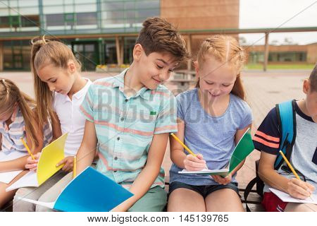 primary education, friendship, childhood, communication and people concept - group of happy elementary school students with notebooks sitting on bench outdoors