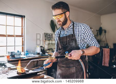 Focused jeweler using  a torch to melt metal in a crucible while working in his jewelry design studio