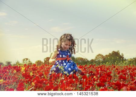 Portrait of adorable smiling brunette girl in blue polka dot dress running in poppy field in sunlight