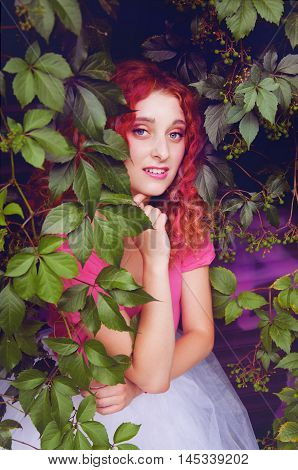 Happy smiling beautiful young woman with curly red hair with colorful hair pieces wearing bouffant skirt and magenta blouse standing among grape leaves