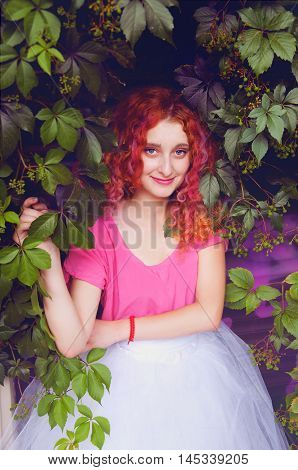Happy smiling beautiful young woman with curly red hair with colorful hair pieces wearing bouffant skirt and magenta blouse standing among grape leaves