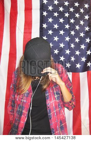 Vintage lifestyle portrait of young hipster girl against american flag background wearing casual clothes black plain t-shirt, red plaid shirt and black snapback cap