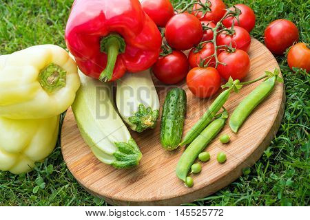 Fresh juicy vegetables on the wooden board on green grass. Top view. Tomato, cucumber, squash, green peas, sweet pepper on the wooden round board on sunny day. Close up. Healthy life concept.
